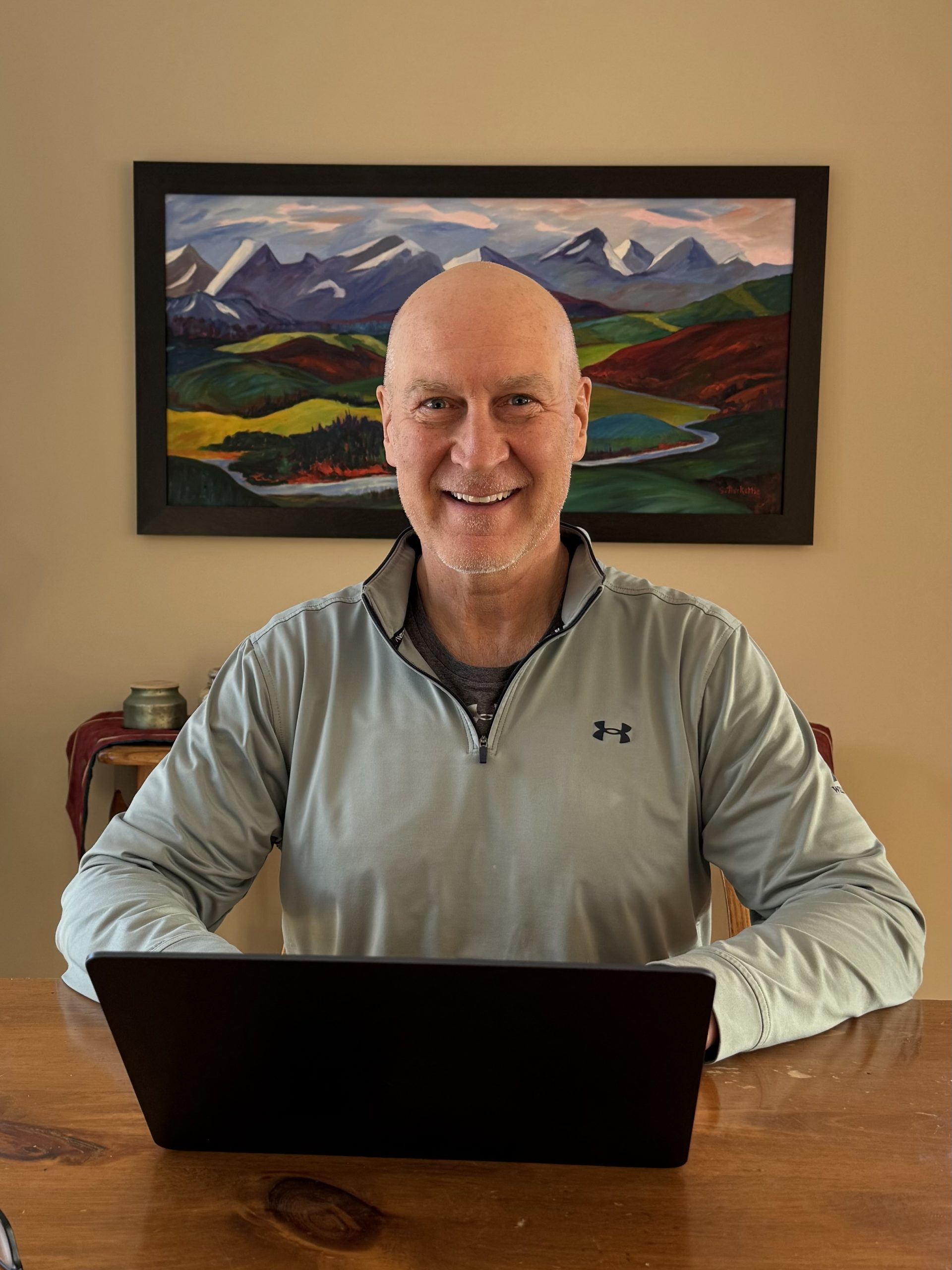 Blogger Rod smiling at a desk with a laptop in front of a mountain landscape painting.