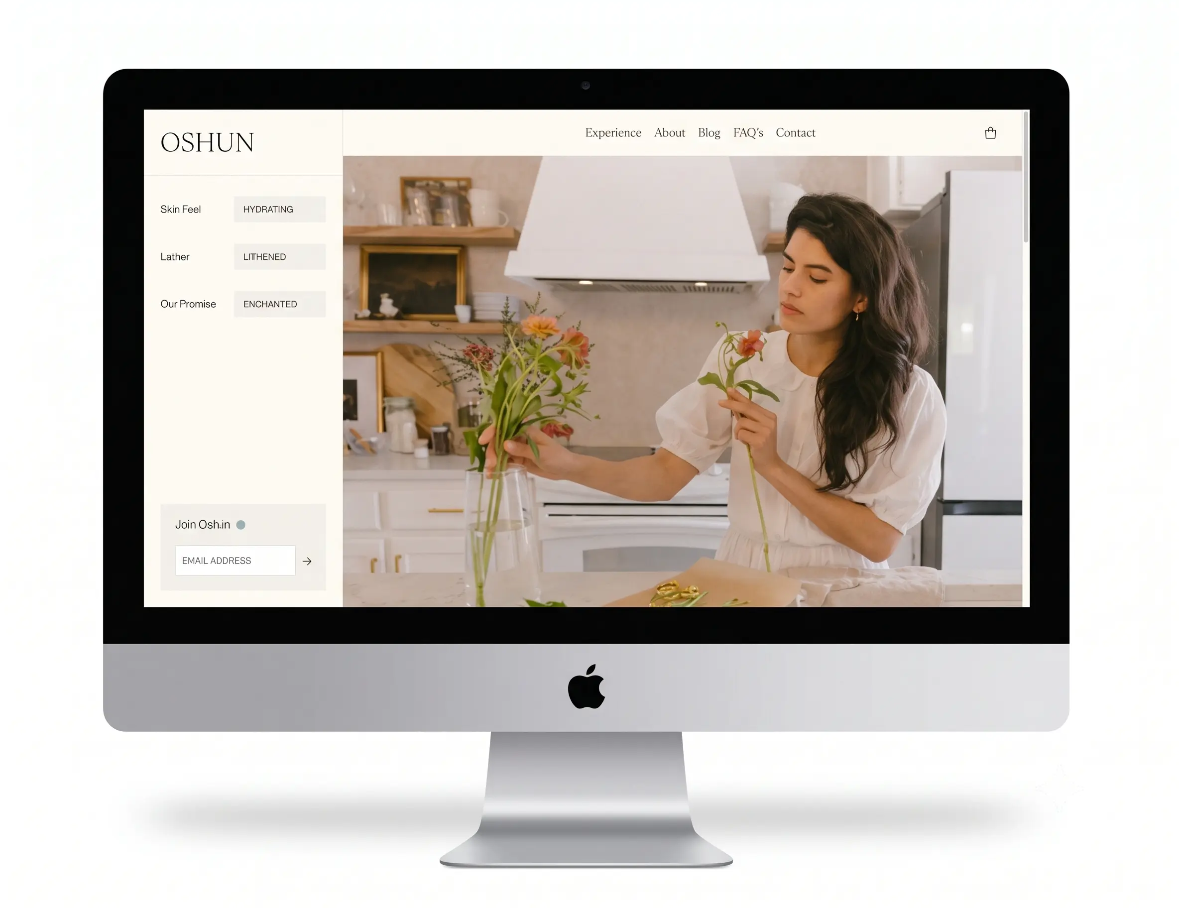 A head-on photograph of a silver Apple iMac computer against a clean white background. The screen displays the 'OSHUN' website, featuring a two-column layout. The left column is beige with the Oshun logo, product attributes (Skin Feel: Hydrating, Lather: Lithened, Our Promise: Enchanted), and an email subscription box. The right column features a lifestyle photograph of a woman arranging flowers in a modern, light-filled kitchen, with a navigation menu (Experience, About, Blog, FAQ's, Contact) across the top.